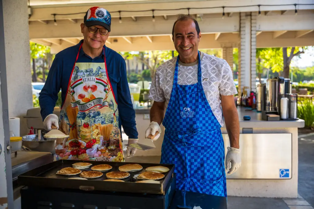 Two men cooking pancakes on a griddle outdoors, both smiling. One wears a blue apron with a decorative hat, the other wears a blue checkered apron. Various cooking utensils and ingredients are visible around them. Background shows trees and kitchen setup.