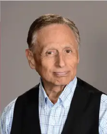 An older man with short gray hair is smiling slightly at the camera. He is wearing a blue plaid button-up shirt and a black vest. The background is a plain gray color.