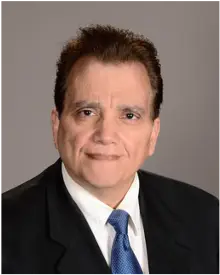 A person with short brown hair wearing a black suit, white shirt, and blue tie, smiling at the camera against a plain gray background.