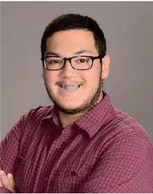 A person wearing glasses and braces smiles while crossing their arms. They are dressed in a maroon plaid shirt against a plain gray background.