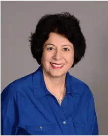 Woman with short black hair wearing a blue collared shirt, smiling against a gray background.