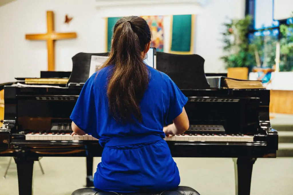 A person with long dark hair in a blue dress plays a grand piano in a church. A wooden cross hangs on the wall, and greenery decorates the room. Sheet music is open on the piano.