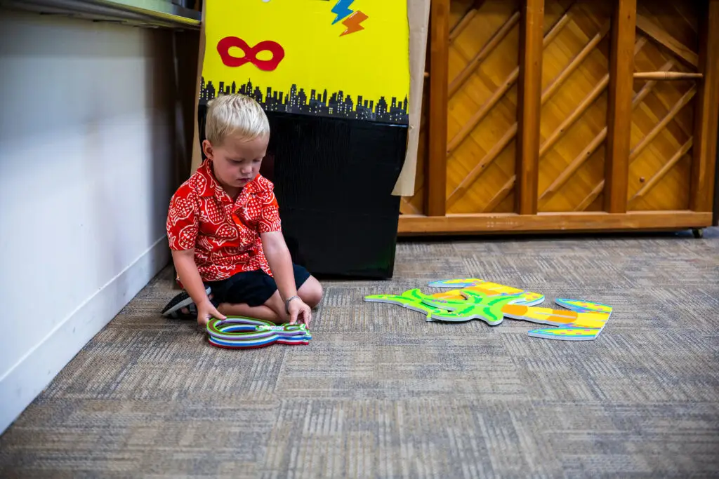 A young child in a red patterned shirt sits on the floor, playing with colorful foam shapes. The background features a wall with a large, yellow superhero-themed decoration and a wooden piano.