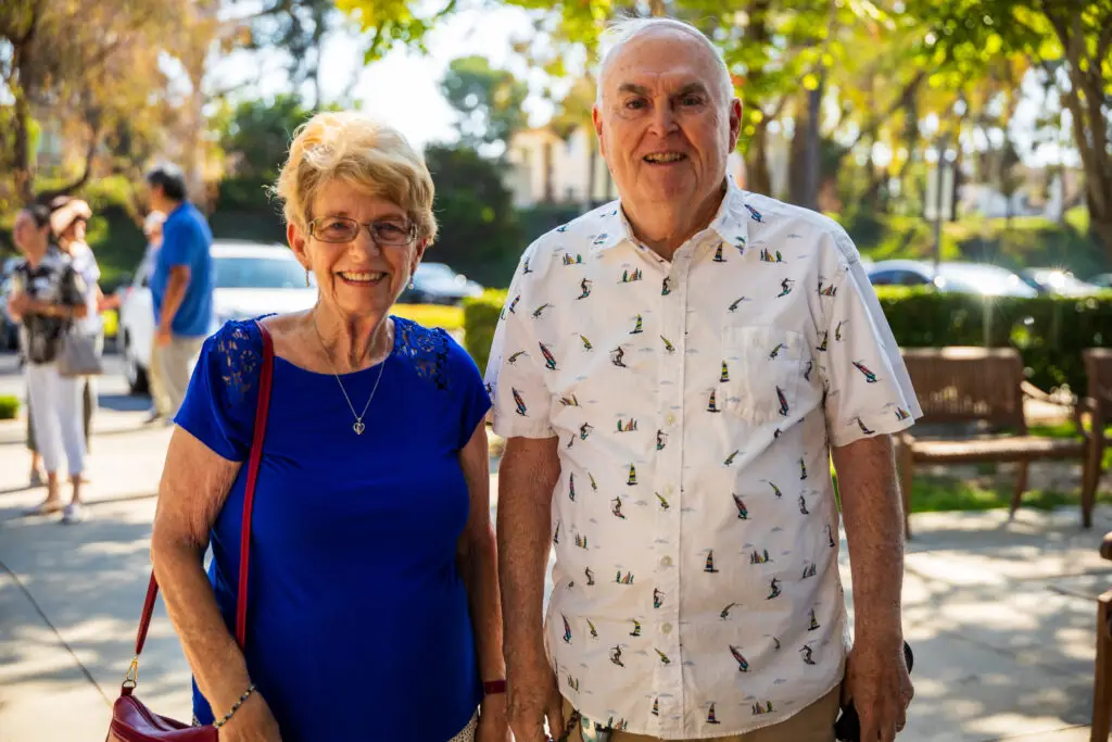 An elderly couple smiling outdoors on a sunny day. The woman wears a blue top and glasses, while the man sports a white shirt with a lighthouse pattern. Trees and people are in the blurred background.