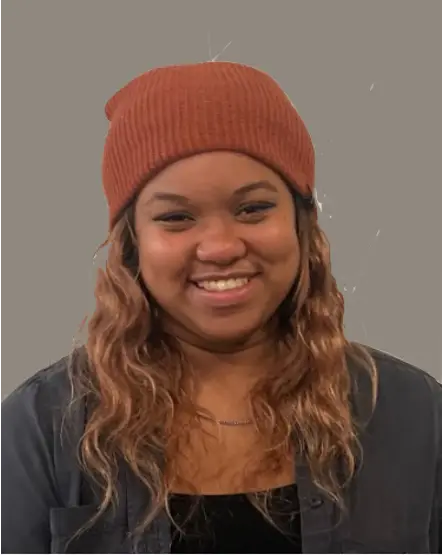 Smiling person wearing a rust-colored beanie and a dark jacket with wavy hair against a plain grey background.