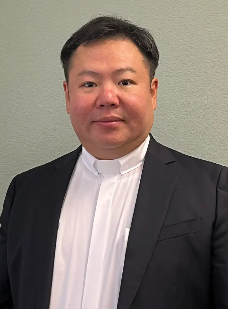 A man in a black suit and white clerical collar stands against a plain light gray background. He has short black hair and a neutral expression.