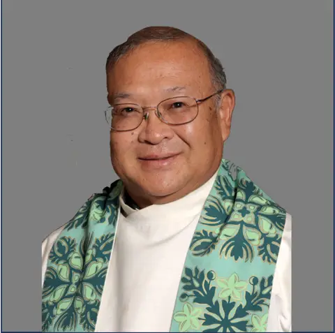 A smiling person wearing glasses and a white robe with a green and teal patterned stole stands in front of a plain gray background.