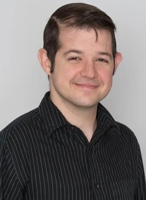 A person with short dark hair smiles at the camera, wearing a black striped shirt against a plain light gray background.