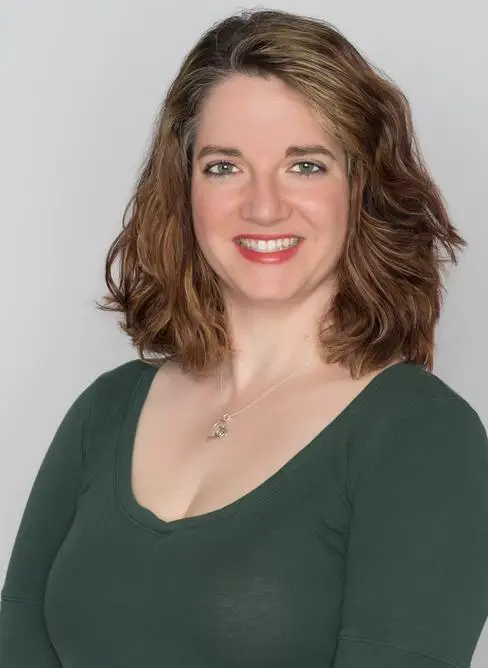 Woman with shoulder-length brown hair and green eyes, smiling, wearing a dark green top and a necklace with a star-shaped pendant, against a light gray background.