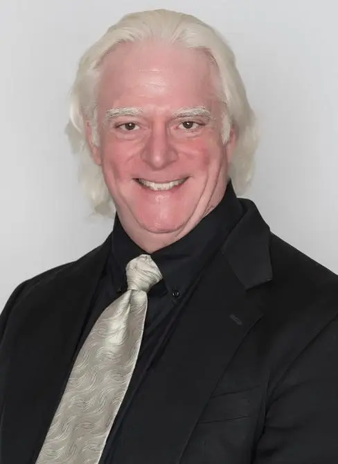 Smiling man with white hair wearing a black suit, black shirt, and silver tie, standing against a plain white background.