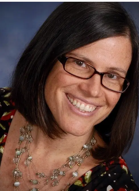 A person with shoulder-length dark hair is smiling at the camera. They are wearing glasses and a necklace with various beads and stones. The background is a gradient of blue tones.