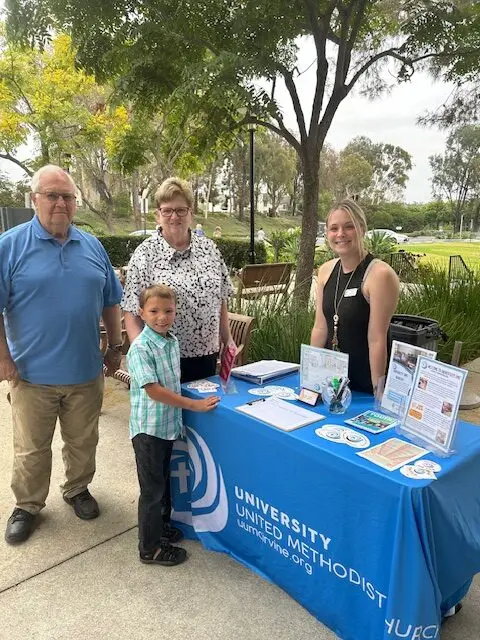 Three adults and one child stand beside a table draped with a University United Methodist Church cover. The table holds brochures and flyers. Trees and a sidewalk are in the background.