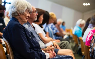 A group of elderly individuals sitting in a row at an indoor event. A woman with white hair and a navy jacket is in focus, sitting in front with eyes closed. Other attendees around her appear attentive. The room has soft lighting and neutral walls.