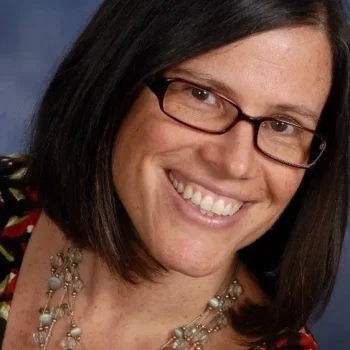 A person with shoulder-length dark hair is smiling at the camera. They are wearing glasses and a necklace with various beads and stones. The background is a gradient of blue tones.